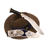 A Khukuri knife with a carbon steel blade and a handle featuring a silver-decorated scabbard with cultural patterns. The knife is displayed with a brown velvet-lined hat and a black bandana, suggesting a traditional or ceremonial use.