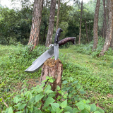 A 10 inch Bajyu Machete Knife with a black leather sheath, hand forged and hunting knife, placed on a wooden stump in a natural outdoor setting.