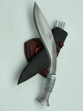 A 12 inch hand forged kukri with a polished blade and a brass handle, accompanied by a black leather scabbard. The knife is displayed on a white background.