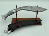 A 12 inch hand forged kukri with a polished blade and a brass handle, accompanied by a black leather scabbard. The knife is displayed on a white background.