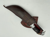A 10 inch hand forged Bowie knife with a black and gray blade and a brown rosewood handle, displayed next to its scabbard.