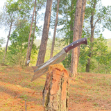 An 11 inch carbon steel handmade cleaver with a wooden handle, resting on a tree stump in a natural outdoor setting for camping and protection.