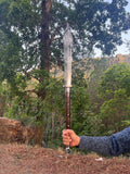 Person holding a machete sword outdoors with trees and mountains in the background