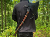 Person in a dark outfit with a large axe over their shoulder in a forest setting